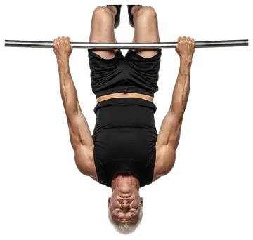 A muscular older man is hanging upside down from a horizontal bar, showcasing his strength. He is wearing a black sleeveless shirt and shorts, with a determined look on his face, emphasizing fitness.