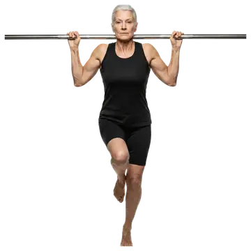 An elderly woman in a black athletic outfit grips a horizontal bar with both hands while balancing on one foot, her other leg hanging down freely. Her expression is focused, showcasing strength and po